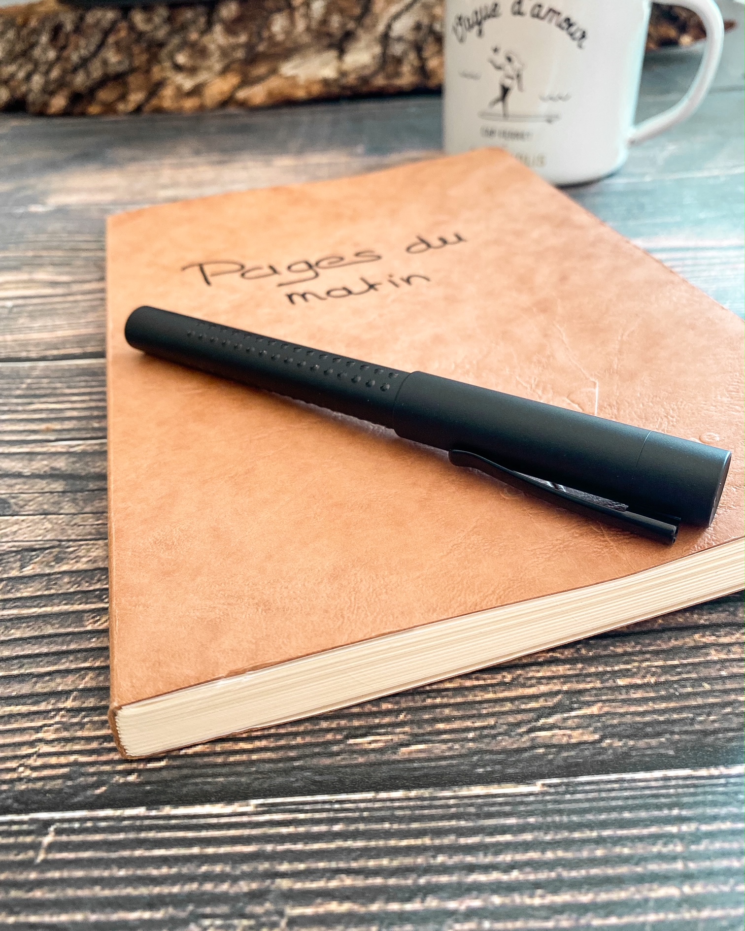 Sur une table en bois, au premier plan un carnet A5 couleur tabac, sur la couverture duquel est écrit à la main "Pages du matin". Un stylo noir est posé sur le carnet. A l'arrière plan, un mug en émail blanc sur lequel est écrit "vague d'amour".