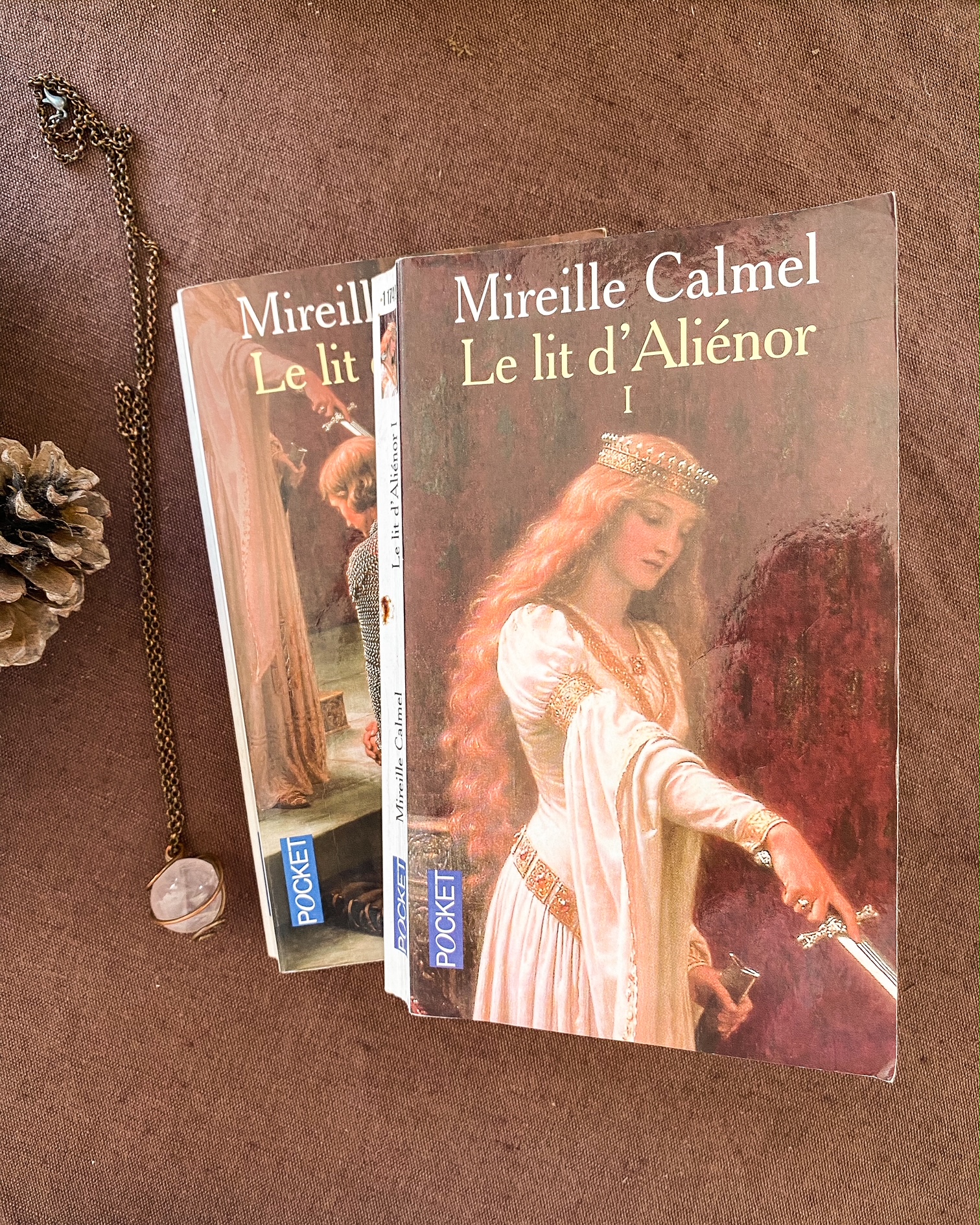 Deux livres de poches posés l'un sur l'autre sur une nape en lin marron. Le livre du dessus représente une partie du tableau "L'accolade" de Blair Leighton avec une femme portant une épée pour adouber un chevalier. A gauche des livres, un pendentif en quartz rose.