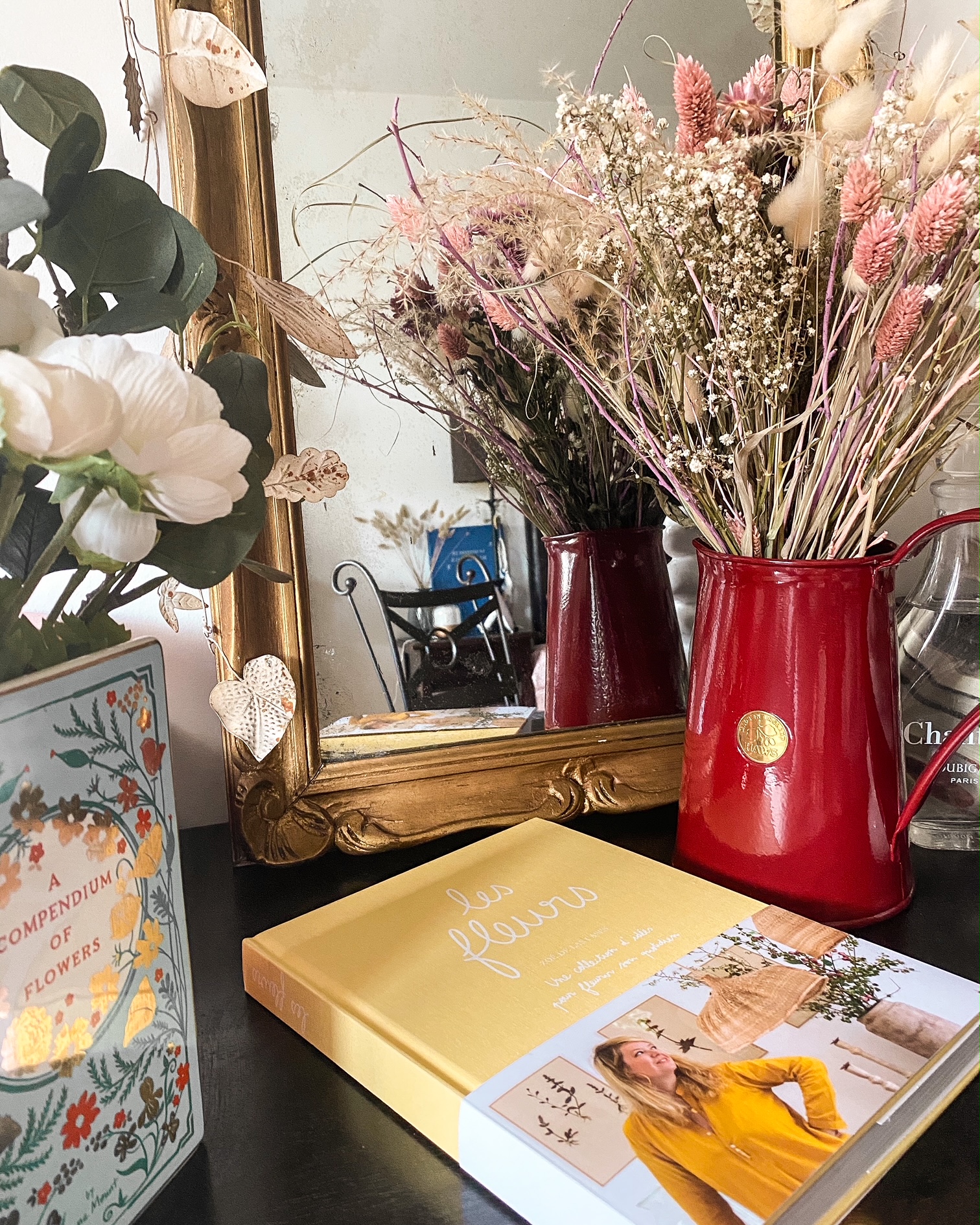 Livre "les fleurs" de Zoé de las cases posé sur une coiffeuse ; un miroir ; un arrosoir rouge avec un bouquet de fleurs séchées ; un vase en forme de livres avec des fleurs blanches et de l'eucalyptus