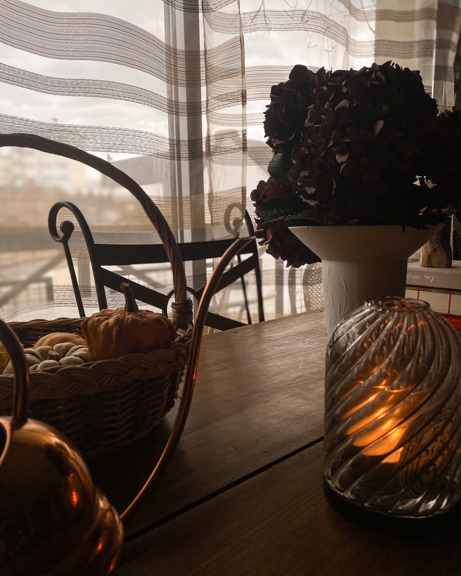 Une table, au fond un vase avec des hortensias, à droite un photophore dans lequel brûle une bougie, à gauche un panier de coloquintes