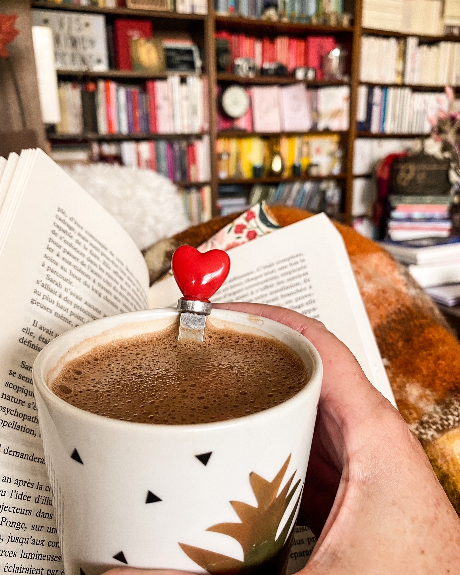 Une tasse de chocolat chaud sur un livre ouvert, un plaid, au fond une bibliothèquett
